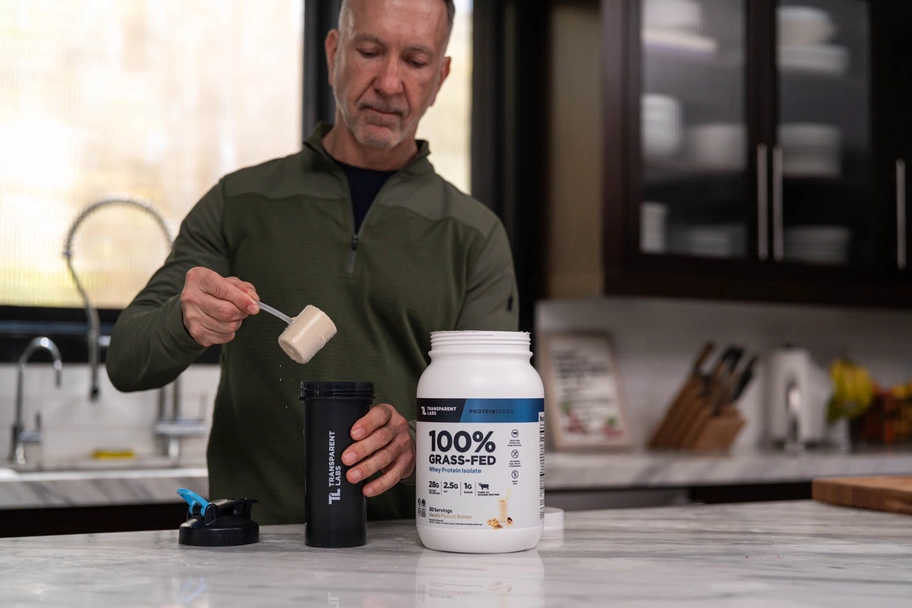 A man scooping whey protein into a shaker bottle at a kitchen counter