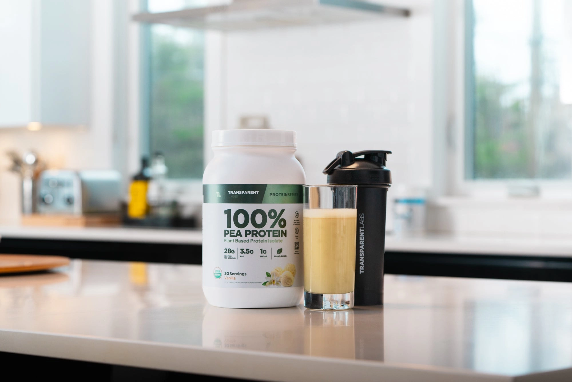 A tub of 100% Grass-Fed whey, a shaker bottle and a glass of smoothie on a kitchen counter
